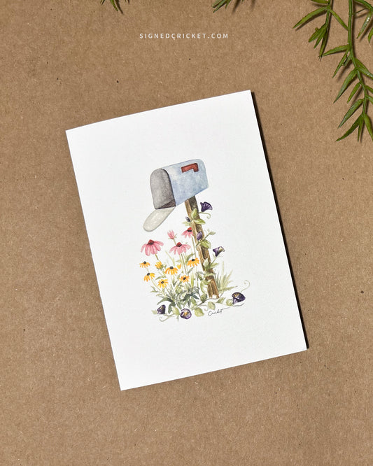 Card featuring a watercolor illustration of a light blue mailbox and wooden post surrounded by 3 types of wildflowers: yellow black-eyed Susans, pink purple coneflowers, and dark purple morning glories. The card is lying flat on a brown paper background.