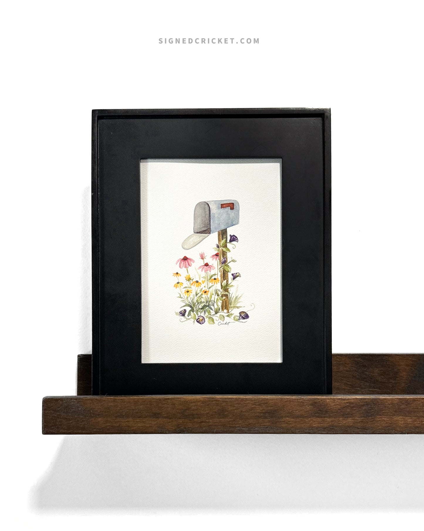 Card featuring a watercolor illustration of a light blue mailbox and wooden post surrounded by 3 types of wildflowers: yellow black-eyed Susans, pinkish purple coneflowers, and dark purple morning glories. The card is framed inside a black minimalistic picture frame on a dark brown wooden shelf against a plain white wall.
