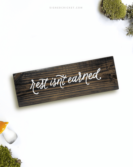 Dark brown wooden sign with 'rest isn't earned' text written on it, lying on a white background