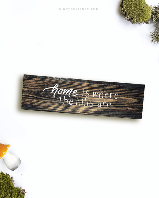 Dark brown wooden sign with 'home is where the hills are' text written on it, lying on a white background