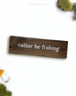 Dark brown wooden sign with 'rather be fishing' text written on it, lying on a white background