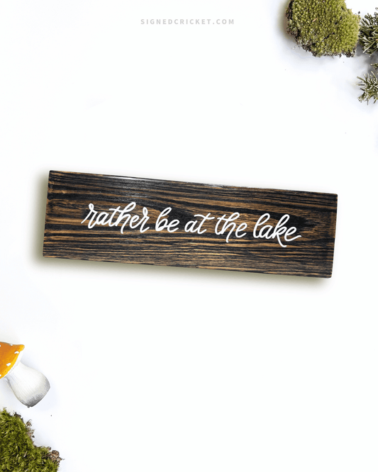 Dark brown wooden sign with 'rather be at the lake' text written on it, lying on a white background