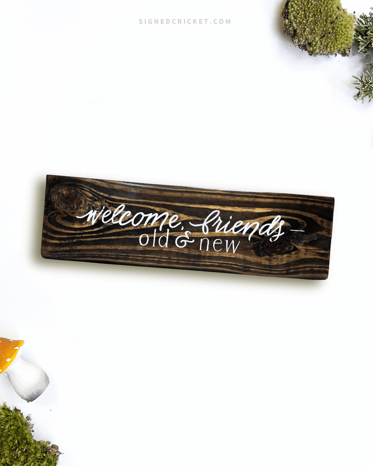 Dark brown wooden sign with 'welcome friends, old and new' text written on it, lying on a white background
