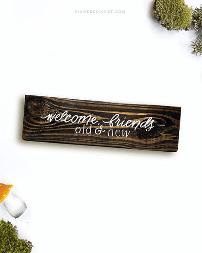 Dark brown wooden sign with 'welcome friends, old and new' text written on it, lying on a white background