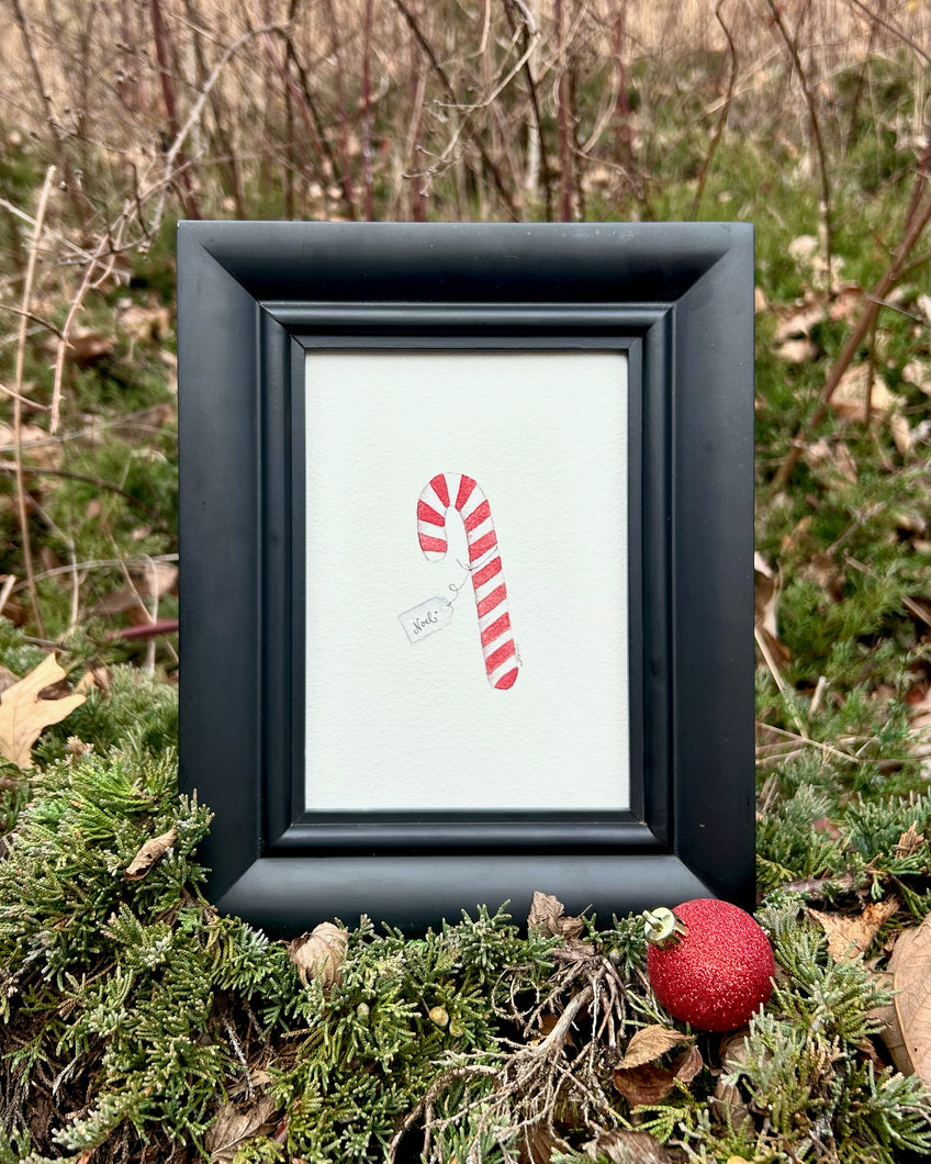 Watercolor painting of a red and white candy cane tied with a handwritten tag that reads “Noel.” Painting is in a beveled black frame. 