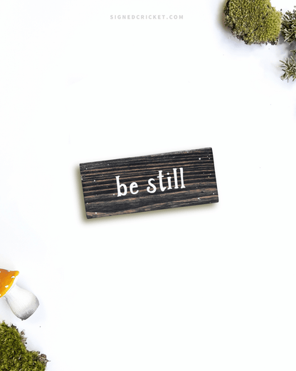 Dark brown wooden sign with 'be still' text written on it, lying on a white background