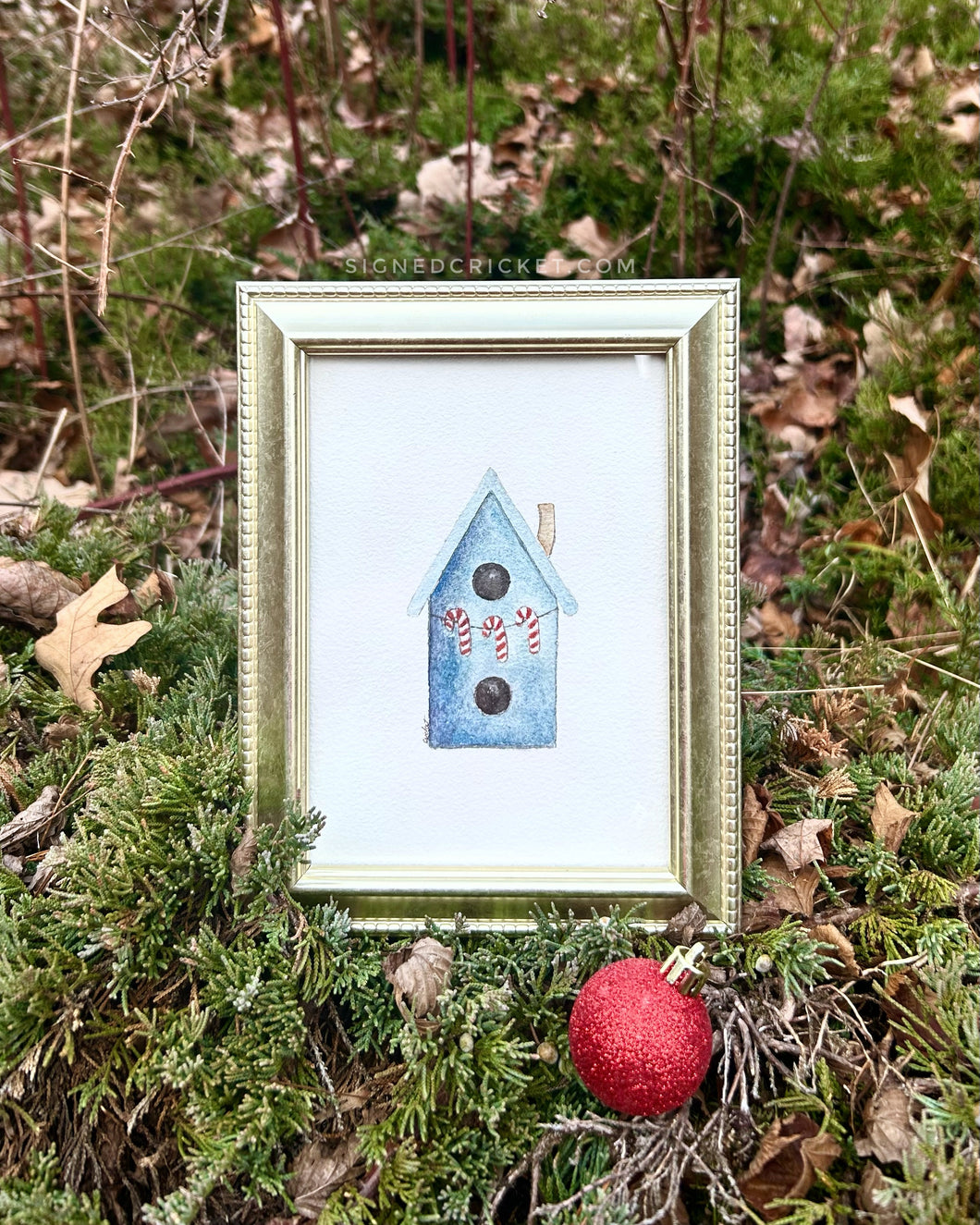 Gold-framed watercolor painting of a blue birdhouse with three red and white candy canes strung across the front.