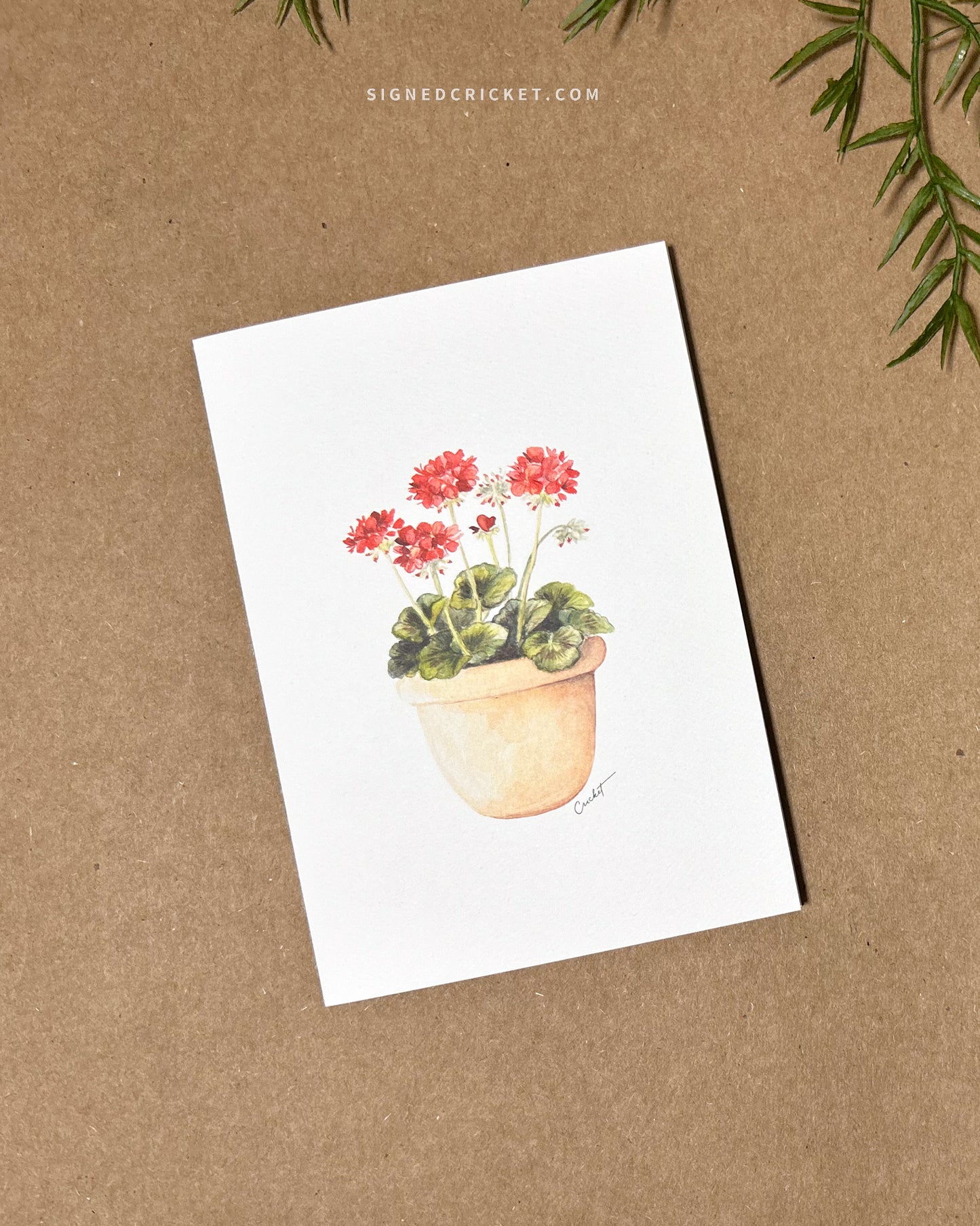 Greeting card featuring a watercolor illustration with a light orange red clay pot with bright red geraniums and dark green leaves against a white background. Card is lying flat on a brown kraft paper background