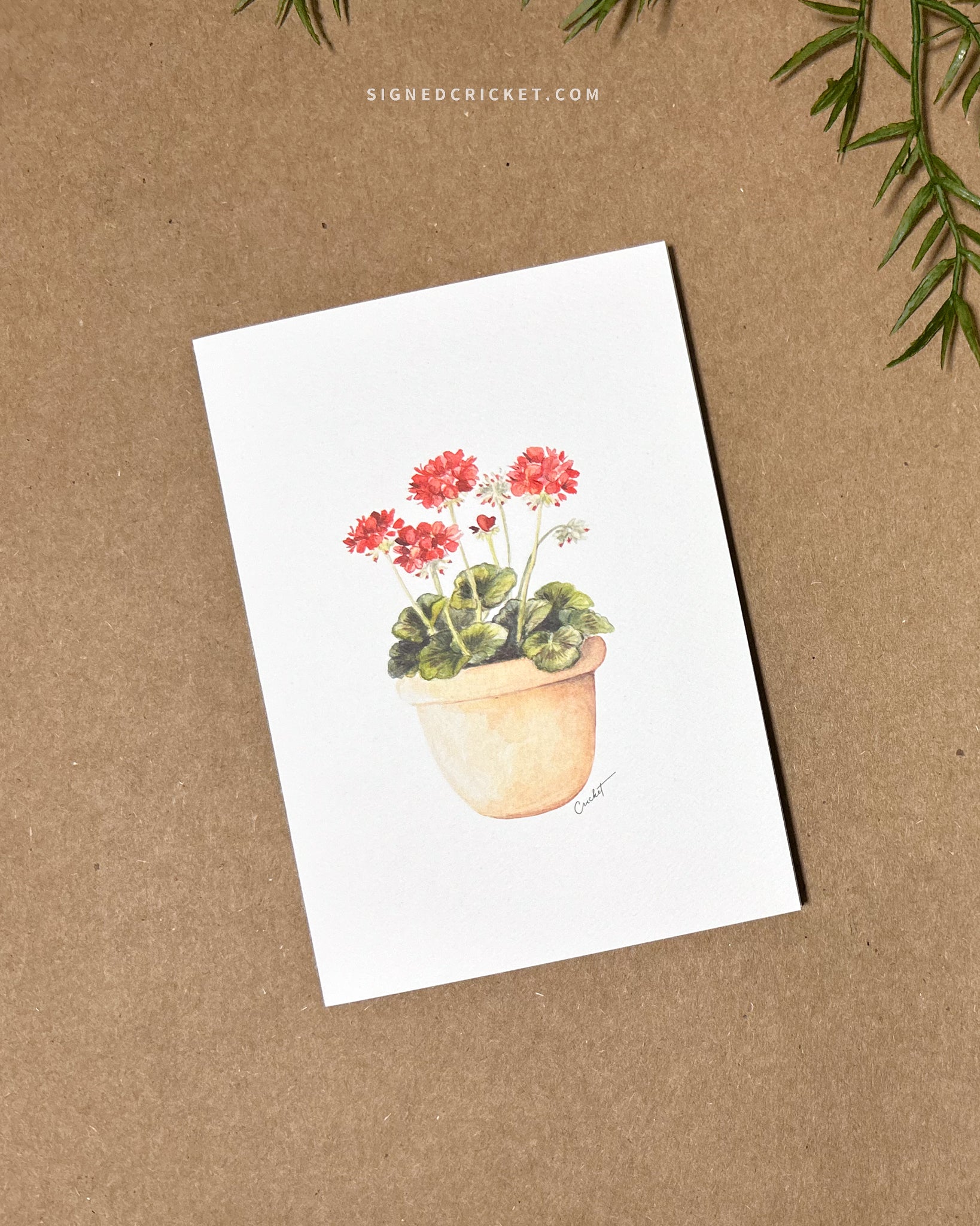 Greeting card featuring a watercolor illustration with a light orange red clay pot with bright red geraniums and dark green leaves against a white background. Card is lying flat on a brown kraft paper background
