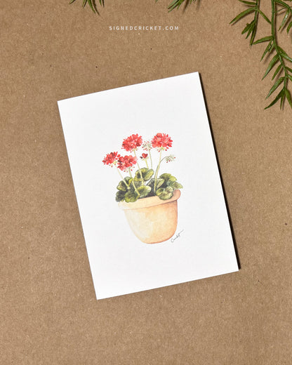 Greeting card featuring a watercolor illustration with a light orange red clay pot with bright red geraniums and dark green leaves against a white background. Card is lying flat on a brown kraft paper background