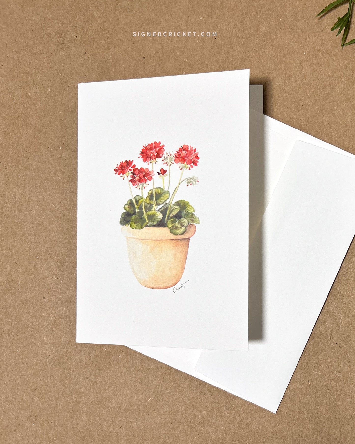 Greeting card featuring a watercolor illustration with a light orange red clay pot with bright red geraniums and dark green leaves against a white background. Card is lying flat on a brown kraft paper background together with a blank white envelope