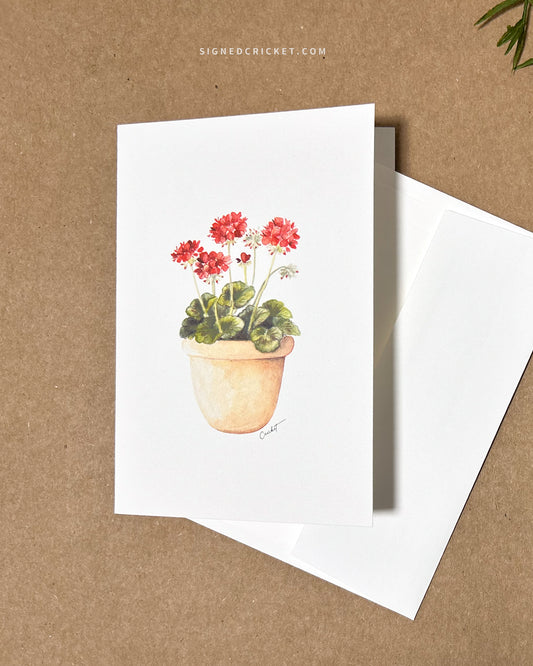 Greeting card featuring a watercolor illustration with a light orange red clay pot with bright red geraniums and dark green leaves against a white background. Card is lying flat on a brown kraft paper background together with a blank white envelope