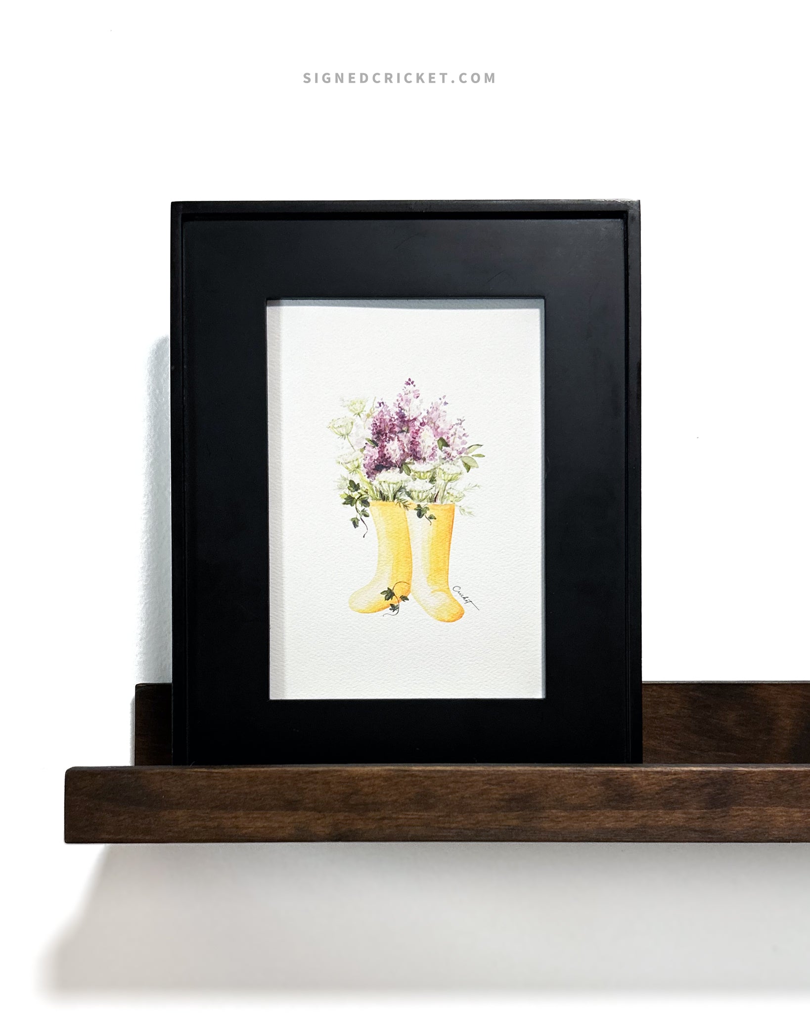 Framed watercolor illustration of yellow rain boots, purple lilacs, green and white Queen Anne's lace, and dark green English ivy inside a wide minimalistic black picture frame on a brown wood floating shelf on a white wall