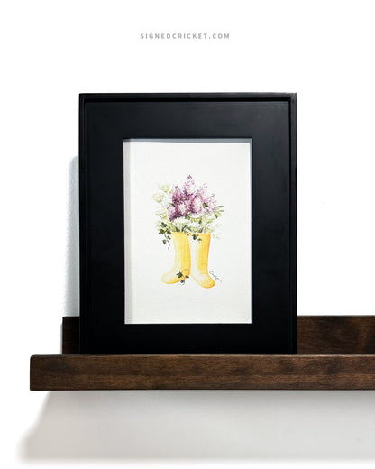 Framed watercolor illustration of yellow rain boots, purple lilacs, green and white Queen Anne's lace, and dark green English ivy inside a wide minimalistic black picture frame on a brown wood floating shelf on a white wall