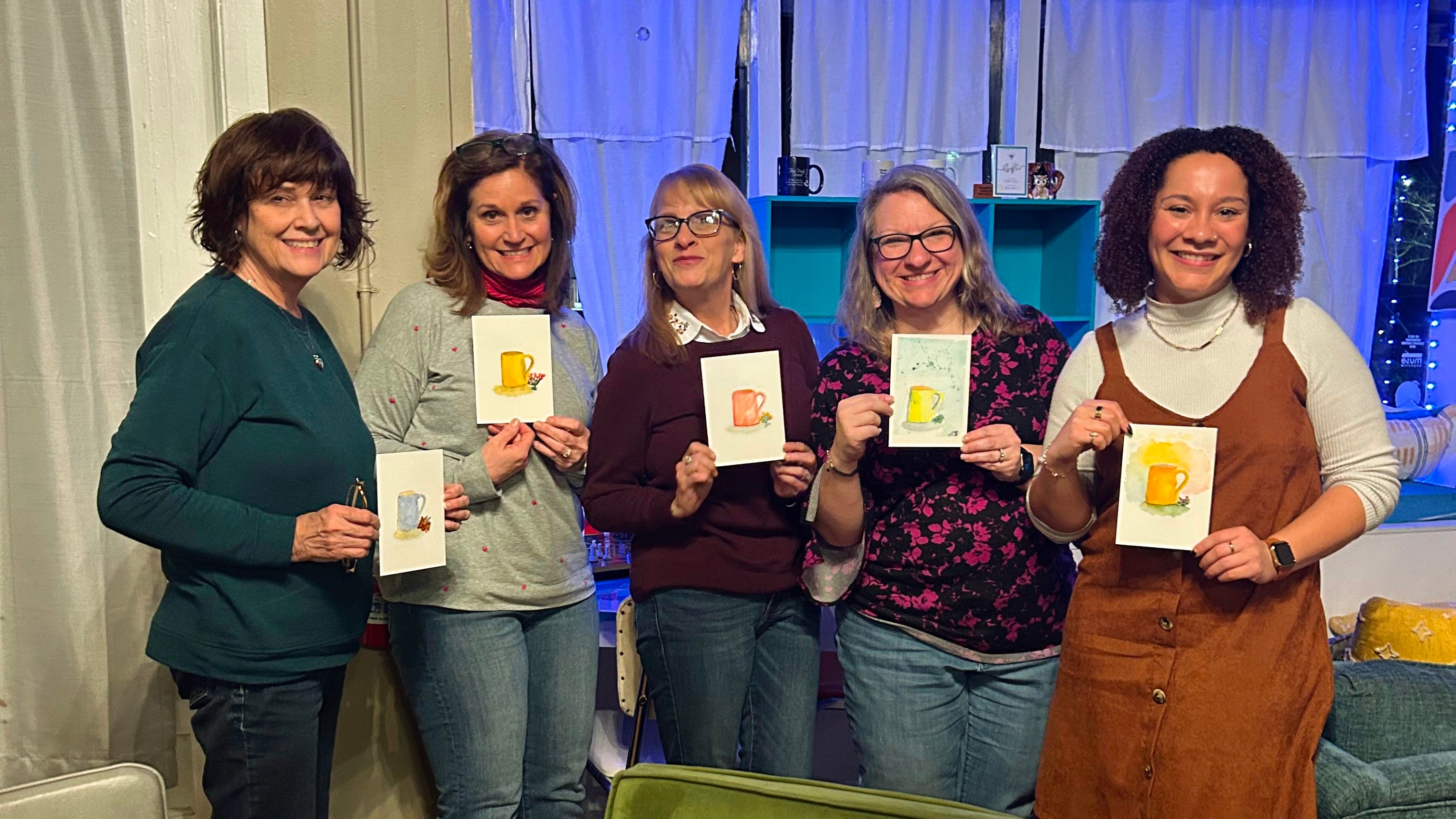 Five smiling women pose holding small paintings of mugs in bright  shades of yellow, red, and blue against a blue curtain backdrop after a Signed Cricket watercolor for beginners workshop