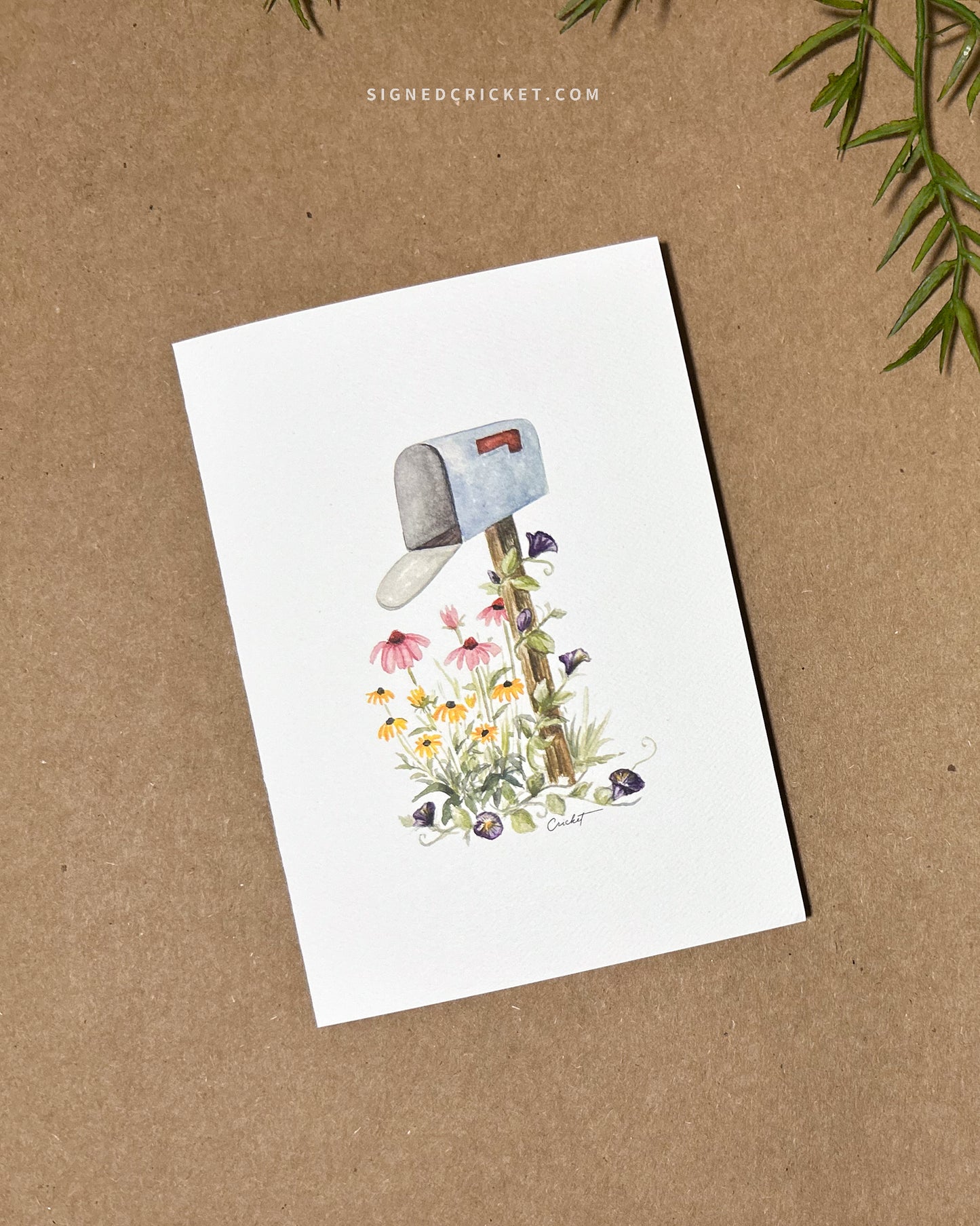 Card featuring a watercolor illustration of a light blue mailbox and wooden post surrounded by 3 types of wildflowers: yellow black-eyed Susans, pink purple coneflowers, and dark purple morning glories. The card is lying flat on a brown paper background.